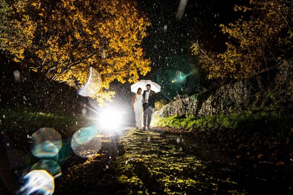 Wedding Photography Best of-121 A couple walks under a white umbrella along a wet, dark path at night, with bright light illuminating them from behind. Autumn trees create a picturesque backdrop, making the scene look like it was captured by a skilled Dodford Manor Wedding Photographer.