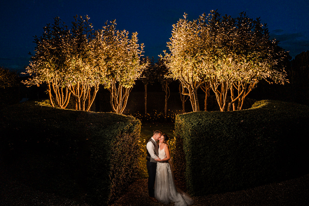 Wedding couple kissing under illuminated trees at night.