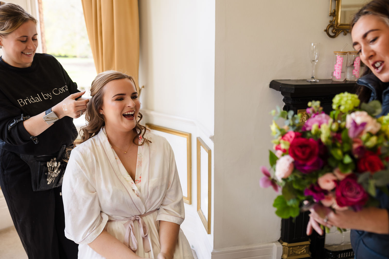 A smiling bride sits in a chair while a hairdresser works on her hair, captured perfectly by Holmewood Hall Wedding Photography. Nearby, another person holds a colorful bouquet of flowers.