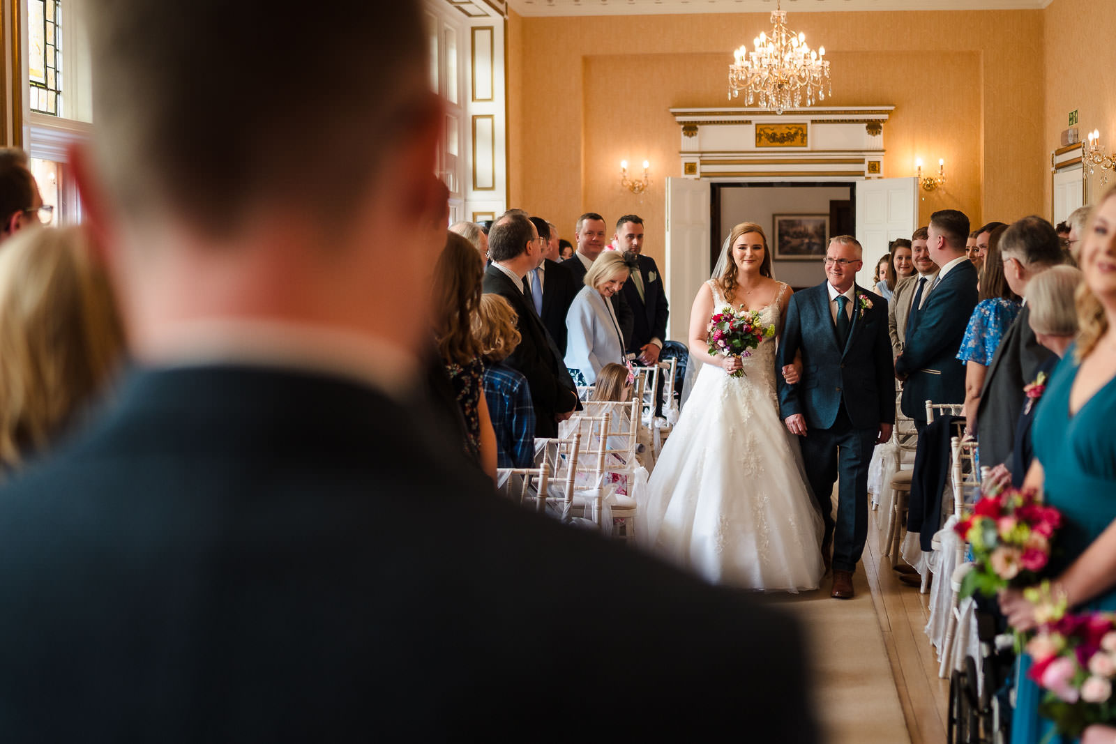Bride in a white gown walks down the aisle with a man in a suit, captured beautifully by Holmewood Hall Wedding Photography, amidst guests seated and standing in an elegantly decorated room with chandeliers.