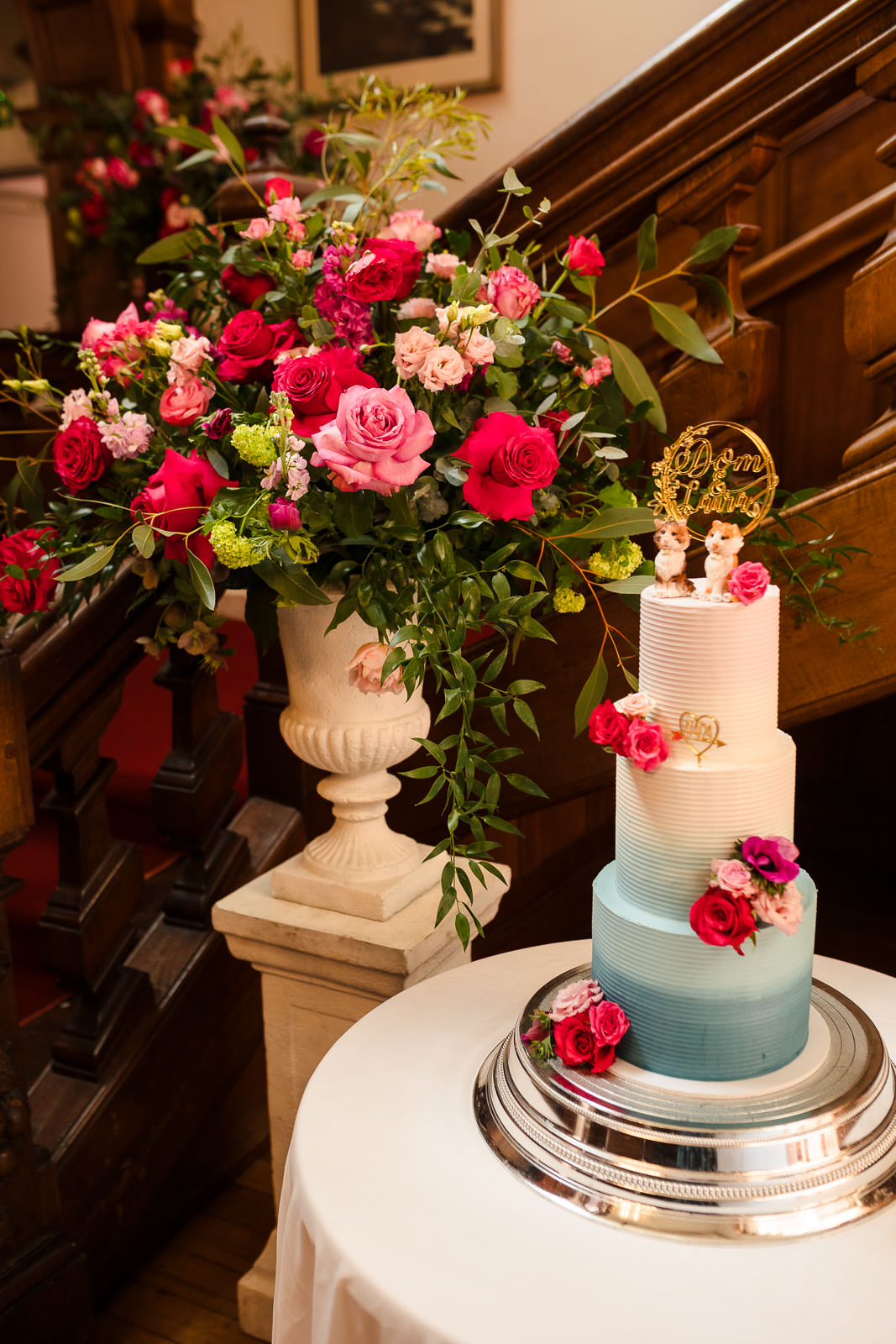 A decorated, three-tiered wedding cake with pink and blue icing, adorned with pink roses, is displayed on a table. A large floral arrangement in a white vase is placed nearby, perfectly captured by Holmewood Hall Wedding Photography.