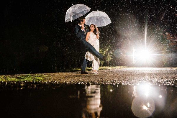 rain wedding photography A couple under clear umbrellas dances on a wet path at night, captured by an all-weather Warwickshire wedding photographer. Raindrops fall as a bright light shines in the background, perfectly reflected in a puddle.