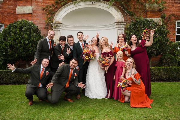 wedding photography portfolio 110 A wedding party poses cheerfully in a garden, expertly captured by a Warwickshire Wedding Photographer. The bride and groom are at the center, surrounded by the bridal party in green and burgundy attire, holding vibrant bouquets.