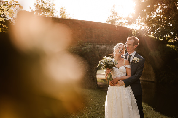 wedding photography portfolio 112 A bride and groom stand together in a garden, smiling and holding a bouquet. Sunlight filters through the trees in the background.