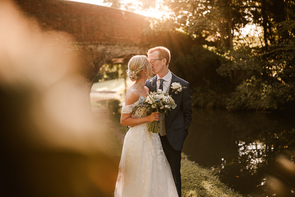 wedding photography portfolio 113 A couple in wedding attire shares a moment by a sunlit riverbank, with a red brick bridge in the background. The bride holds a bouquet, both dressed formally. Captured beautifully by a Warwickshire wedding photographer, their love story is etched into this picturesque scene.