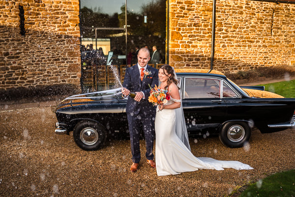 wedding photography portfolio 125 A smiling bride and groom stand in front of a classic car. The groom opens a bottle of champagne, causing it to spray. They are outdoors, with a stone building in the background.