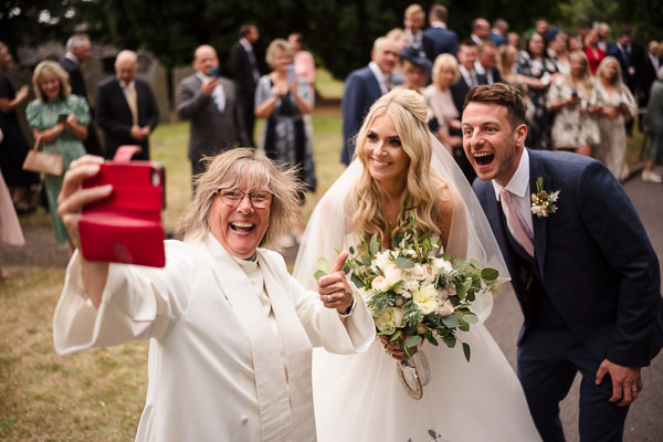 wedding photography portfolio 13 A Warwickshire wedding photographer captures a joyful moment as the bride and groom take a selfie with the thumbs-up officiant, surrounded by their cheering guests at the outdoor ceremony.