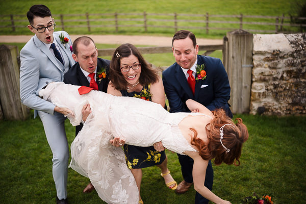 wedding photography portfolio 21 A group of four people playfully lift a bride in a white dress on a grassy area, with wooden fencing and a stone wall as the perfect backdrop for this picturesque moment captured by your Warwickshire wedding photographer.
