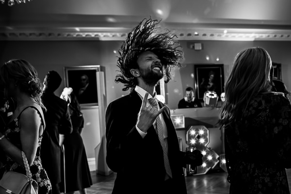 wedding photography portfolio 22 A person in formal attire enthusiastically dancing, with long hair flying, at a lively indoor event. Others are also present, and the room is dimly lit with highlighted portraits on the walls.