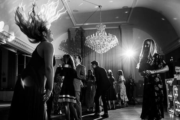 wedding photography portfolio 23 A black and white photo of people dancing in a room with a chandelier. A person in the foreground with long hair is flipping it back, while others dance in the background.