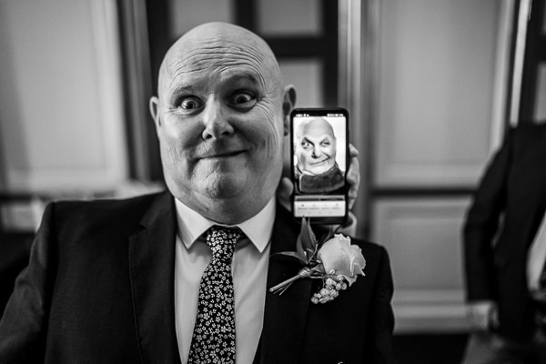 wedding photography portfolio 28 A grinning man in a suit holds a phone displaying a photo of himself making a similar expression. Black and white image.