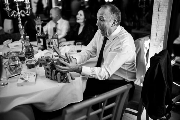 wedding photography portfolio 29 A man in a suit is seated at a table, reacting excitedly as wooden blocks tumble in a game of Jenga. Captured with the precision of a Warwickshire Wedding Photographer, the scene buzzes with life, surrounded by other guests and various delightful details around the table.