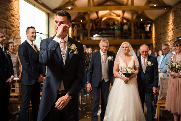 wedding photography portfolio 41 A groom wipes his eye as the bride walks down the aisle, accompanied by two men in a rustic wedding venue. Guests are seated on wooden chairs, and the atmosphere is emotional.