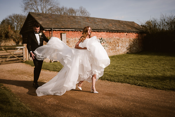 wedding photography portfolio 66 A bride with a flowing white dress walks on a dirt path, holding flowers, beside a groom in a black suit. A rustic building and greenery are in the background.