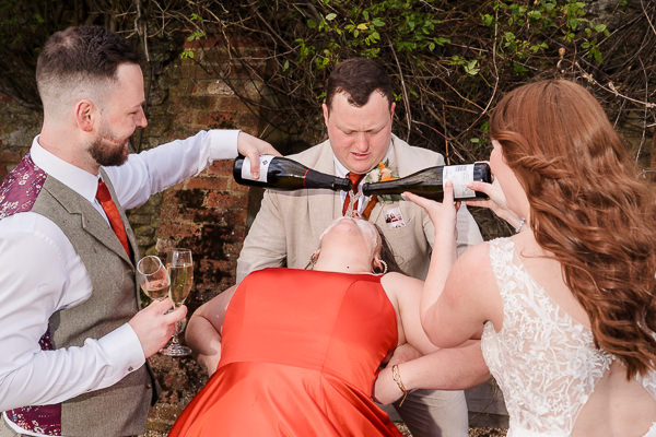 wedding photography portfolio 71 A group celebrates outdoors. A woman in a red dress leans back while two people pour drinks from bottles into her mouth. Two others hold her arms and a glass of champagne.