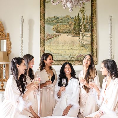 wedding photography portfolio 80 Six women in robes sit on a bed in a bright room with a chandelier, holding glasses of champagne and smiling. A tapestry depicting a scenic landscape hangs on the wall behind them.