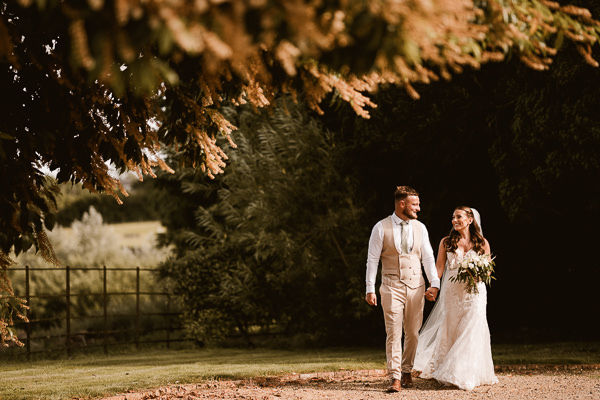 wedding photography portfolio 82 A bride and groom holding hands and smiling walk on a path in a garden setting, surrounded by trees.