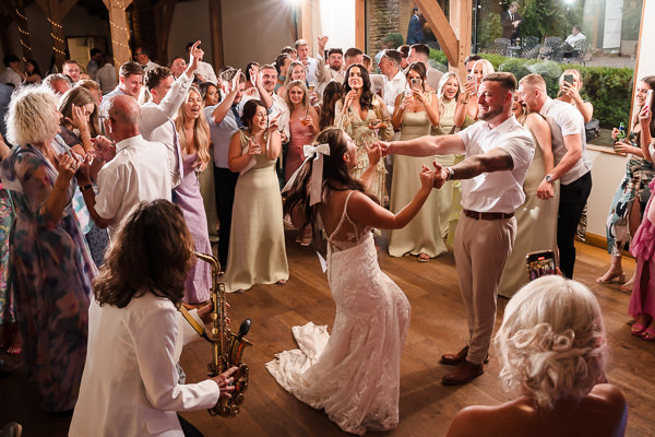 wedding photography portfolio 84 A bride and groom dance joyfully at a wedding reception, surrounded by cheering guests. A saxophonist plays in the foreground.