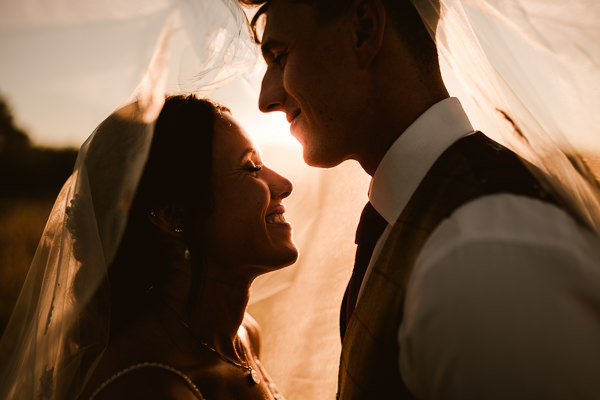 wedding photography portfolio 85 A bride and groom stand close, smiling under a veil with sunlight behind them.