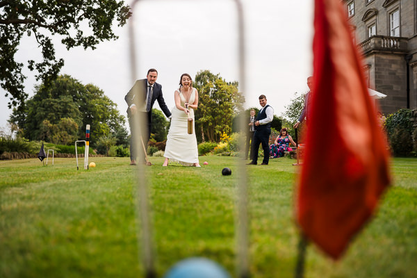 wedding photography portfolio 99 A bride and groom play croquet on a lush lawn, the bride gracefully swinging the mallet. Guests watch with delight as a Warwickshire Wedding Photographer captures the moment. A vibrant red flag and croquet hoops add charm to this picturesque scene.