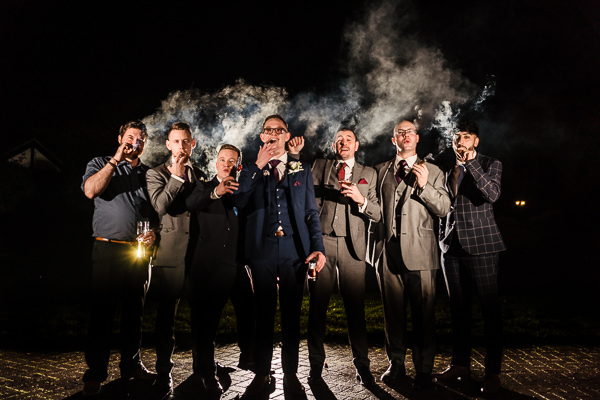 winter low light wedding photographer 10 Seven men in suits stand in a row at night, smoking cigars with smoke in the background.
