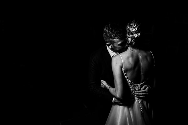 winter low light wedding photographer 20 A bride and groom embrace closely in a dimly lit setting, with the bride's back facing the camera.