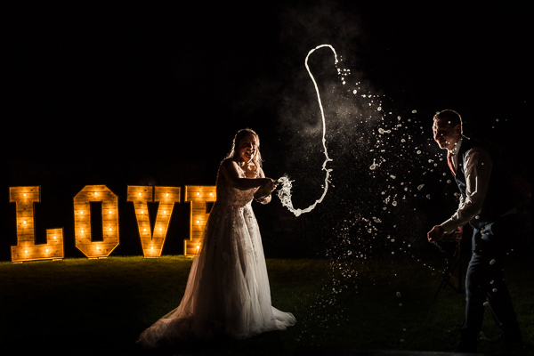 winter low light wedding photographer 38 A bride and groom joyfully popping a champagne bottle at night with illuminated "LOVE" letters in the background.