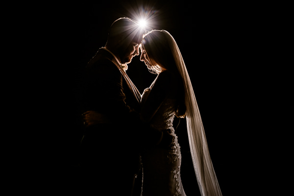winter low light wedding photographer 39 A silhouetted couple in wedding attire stands close together with a bright light behind them, creating a halo effect—a magical moment captured by Winter Wedding Photographers Warwickshire.