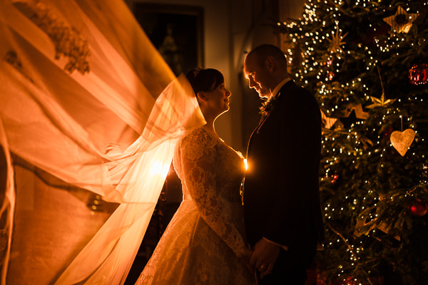 winter low light wedding photographer 41 A bride and groom stand facing each other beside a decorated Christmas tree, captured beautifully by a Milton Keynes wedding photographer. The bride's veil is illuminated, creating a dramatic light effect.