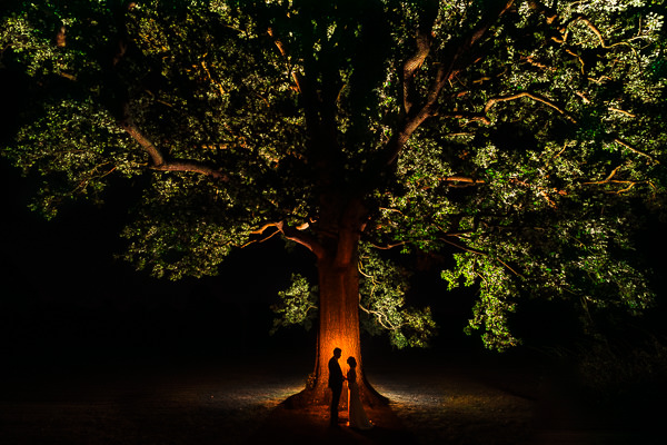 winter low light wedding photographer 45 A large tree at night with its branches and leaves illuminated creates a magical scene fit for Milton Keynes Winter Wedding Photography. Two silhouetted figures stand beneath it, adding an air of mystery to the enchanting moment.