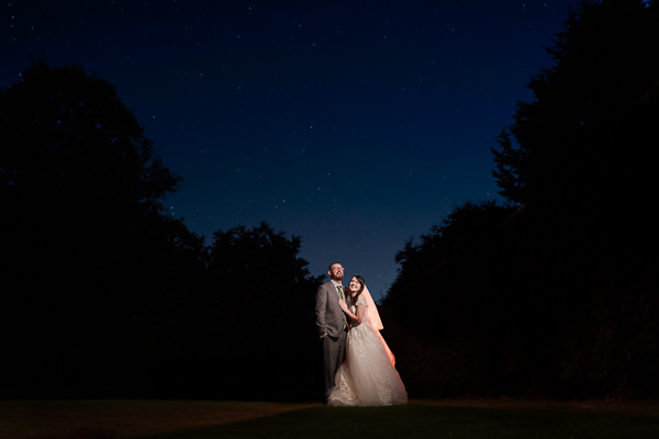 winter low light wedding photographer 46 A bride and groom stand together under a starlit sky, captured beautifully by a Milton Keynes wedding photographer, surrounded by silhouettes of trees.