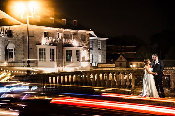 winter low light wedding photographer 47 A couple embraces on a bridge at night, with blurred lights from passing cars in the foreground. A lit building is in the background.