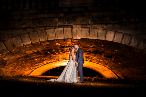winter low light wedding photographer 48 A bride and groom stand under a stone archway at night, illuminated by warm light, beautifully captured by a Milton Keynes wedding photographer.