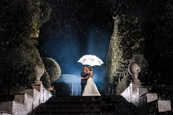 winter low light wedding photographer 49 A couple stands under a white umbrella on stone steps in a garden at night, captured by renowned winter wedding photographers in Warwickshire. They are surrounded by rain, with a building visible in the background.