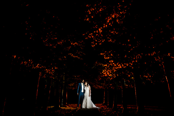 winter low light wedding photographer 7 Bride and groom stand on a path under trees with autumn leaves, illuminated dramatically against a dark background.