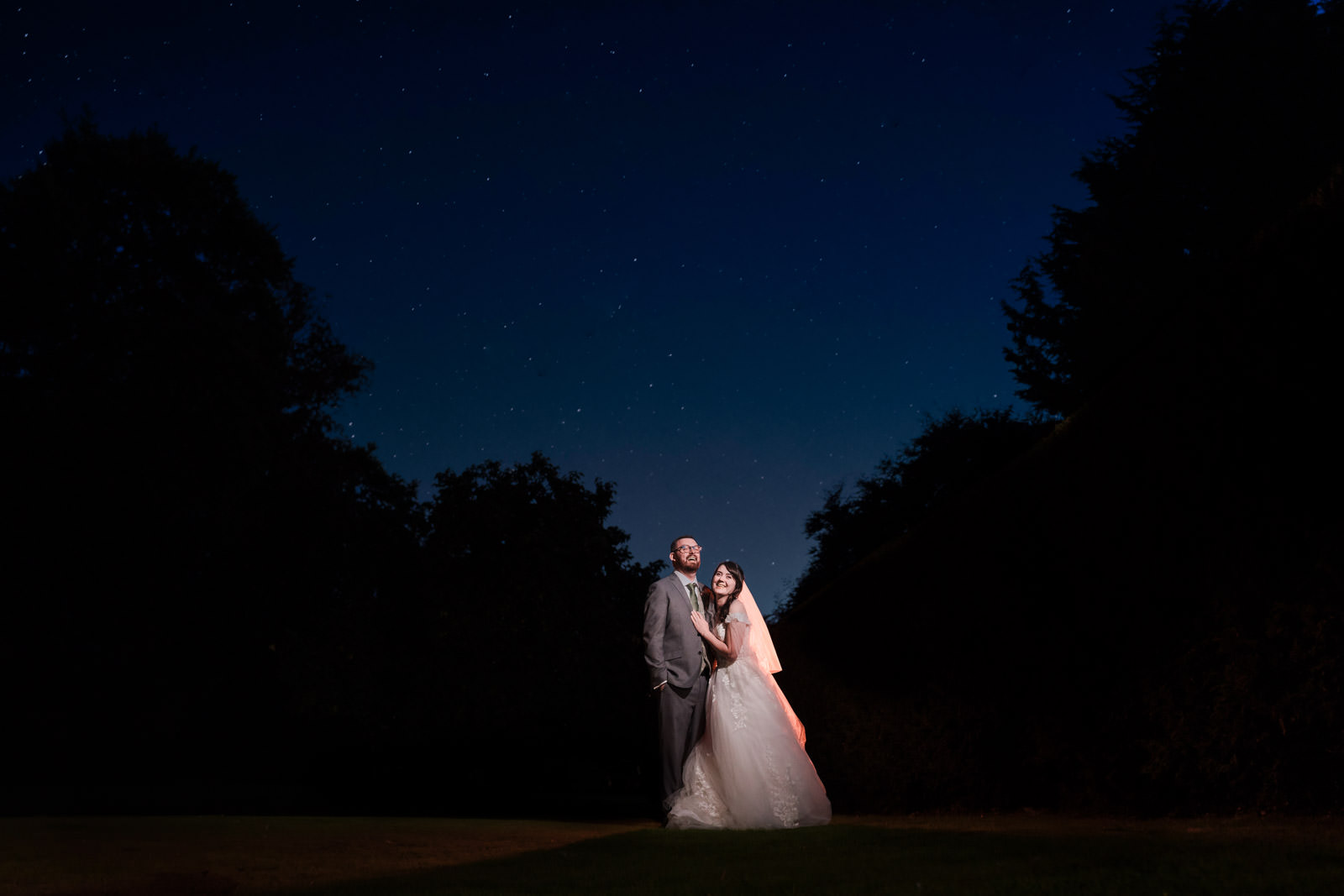 A bride and groom stand outdoors at night under a starry sky, illuminated by soft lighting, with trees silhouetted in the background.