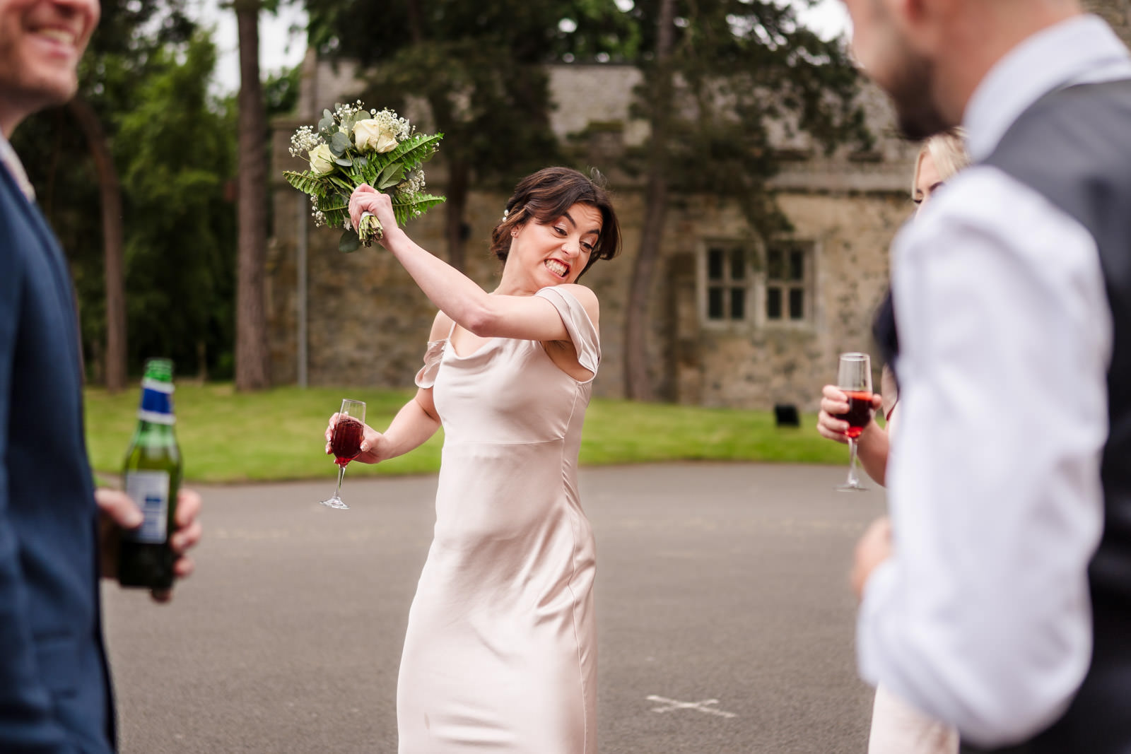 A woman in a light dress playfully swings a bouquet, surrounded by people holding drinks at an outdoor event.
