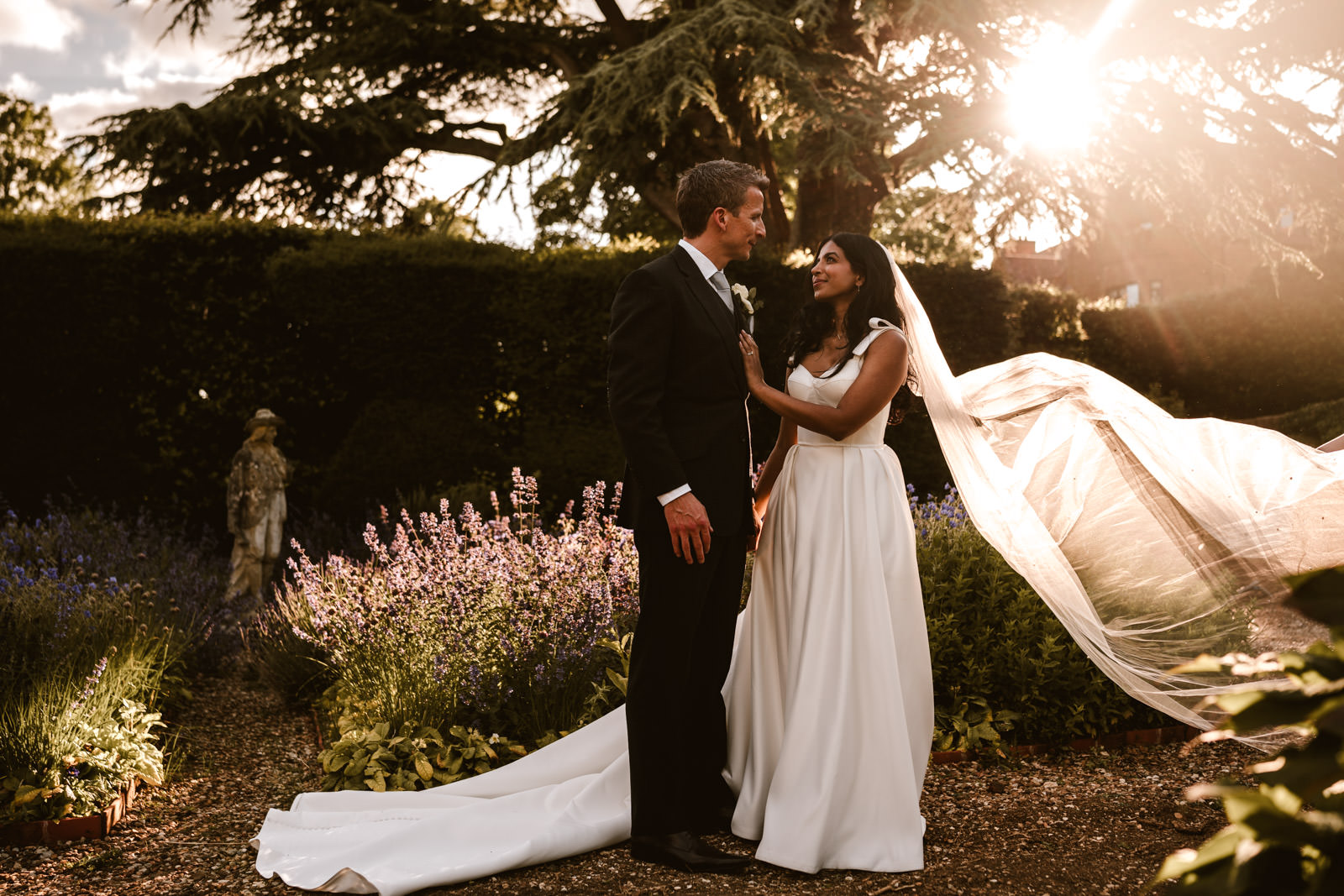 A bride and groom stand together in a garden at sunset, with the bride’s veil flowing behind her and sunlight streaming through the trees.
