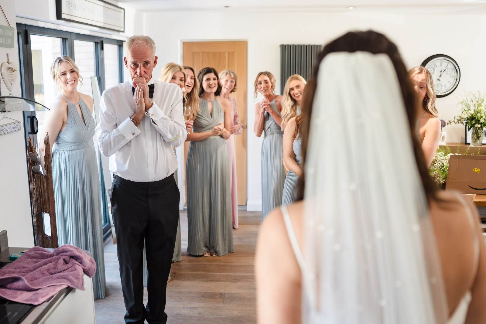 A man reacts emotionally as he sees a bride in a white dress and veil, surrounded by bridesmaids in light blue dresses inside a bright room.