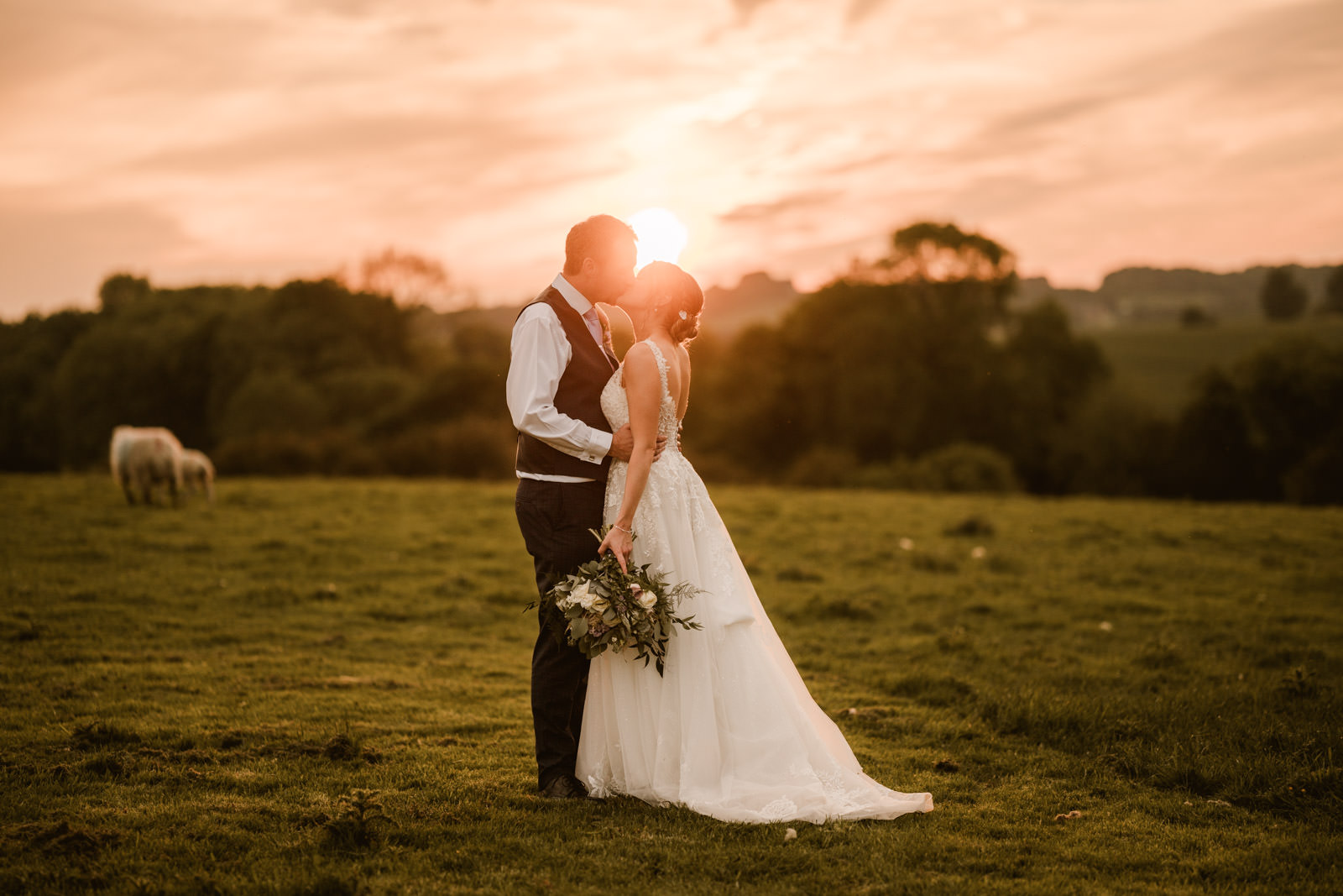 A bride and groom stand in a grassy field at sunset, holding a bouquet and embracing, with trees and a grazing animal in the background.