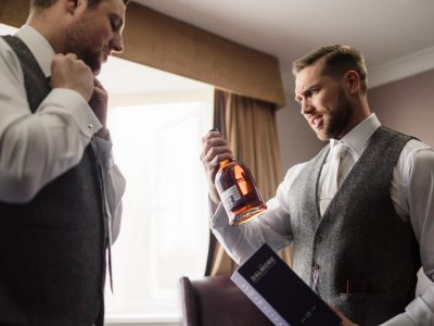 benefits of two photographers 12 Two men in suits are preparing for an event, captured in a groom prep photography session. One adjusts his tie while the other, potentially part of a husband and wife photography team, holds a bottle of whiskey and a box, carefully examining the label.