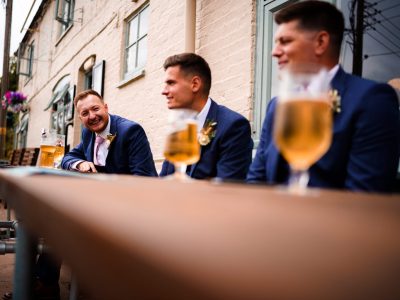 benefits of two photographers 15 Three men in blue suits sit at an outdoor table with drinks, smiling and talking about the benefits of two wedding photographers, near a brick building.
