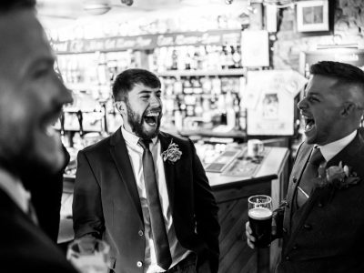 benefits of two photographers 16 Three men in suits are laughing together, holding drinks in a lively bar setting. As conversations flow like fine wine, they discuss if hiring husband and wife photographers for a unique perspective could add charm to future events.