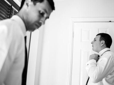 benefits of two photographers 19 Two men in dress shirts and ties, one adjusting his tie while the other looks away. The black and white image, likely captured by husband and wife photographers, exudes timeless elegance in a room with a door subtly framing the scene.