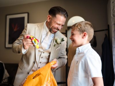 benefits of two photographers 25 Man in a beige suit smiling at a boy while holding a colorful item from an orange bag. The boy looks up, grinning. A framed picture and curtain provide the backdrop, capturing the enchanting moment that husband and wife photographers expertly immortalize together.