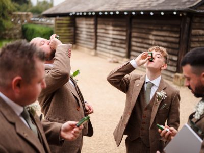 benefits of two photographers 27 Four men in brown suits take shots from small bottles outdoors near a wooden building, capturing the essence of groom prep photography.