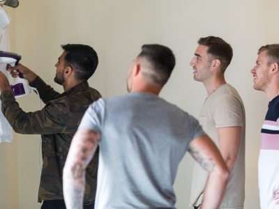 benefits of two photographers 4 Four men stand in a room. One uses a handheld steamer on a white shirt while the others watch, highlighting the intricate moments often captured during groom prep photography.