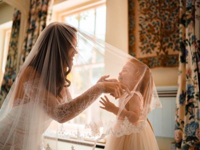 bride getting ready 12 A bride getting ready, playfully interacts with a young girl in a dress near a sunlit window. Two photographers capture the moment, as floral tapestries adorn the walls and the second shooter captures the joy surrounding.