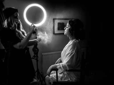 bride getting ready 15 In a dimly lit room, under the soft glow of a ring light, a bride getting ready sits serenely in her robe while a second shooter captures the moment as another woman applies spray.
