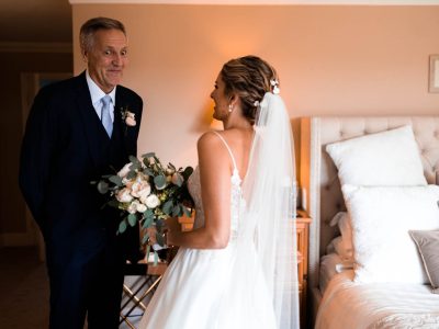 bride getting ready 18 In a warmly lit bedroom, the bride holds a bouquet and smiles at an older man in a suit with a playful expression as the second shooter captures these cherished moments.